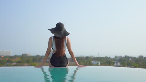 a woman perched on the edge of a resort pool sits with her back to the camera as she looks over the cityscape beyond