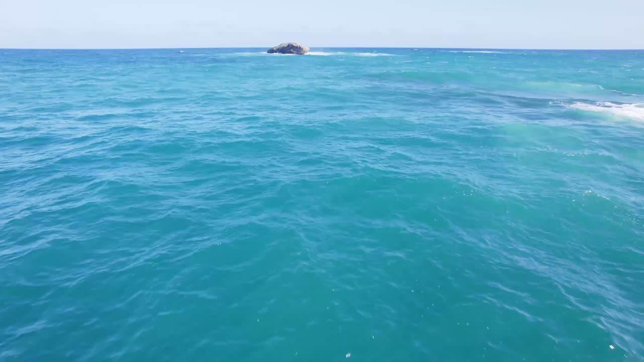 Crystal-clear blue ocean stretches to the horizon with gentle waves and a small rocky island in the distance under a bright sunny sky.