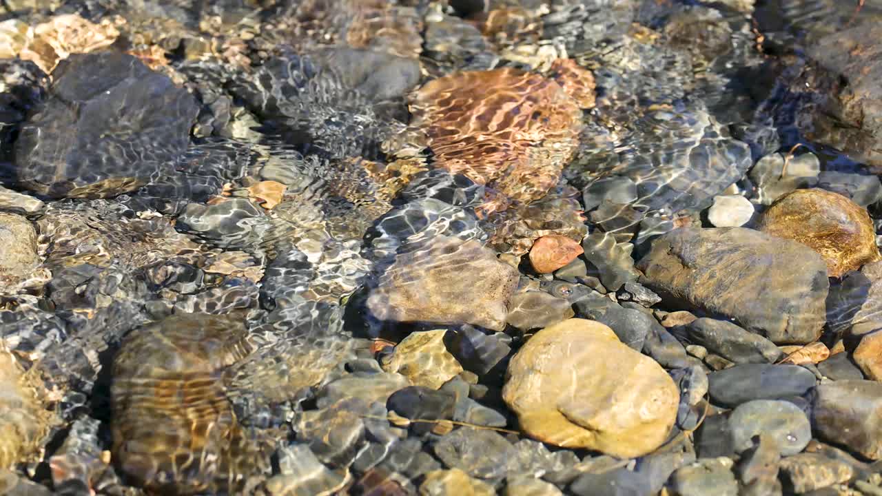 Sunlit creek water flows over multicolored stones, creating ripples and reflections, static camera angle