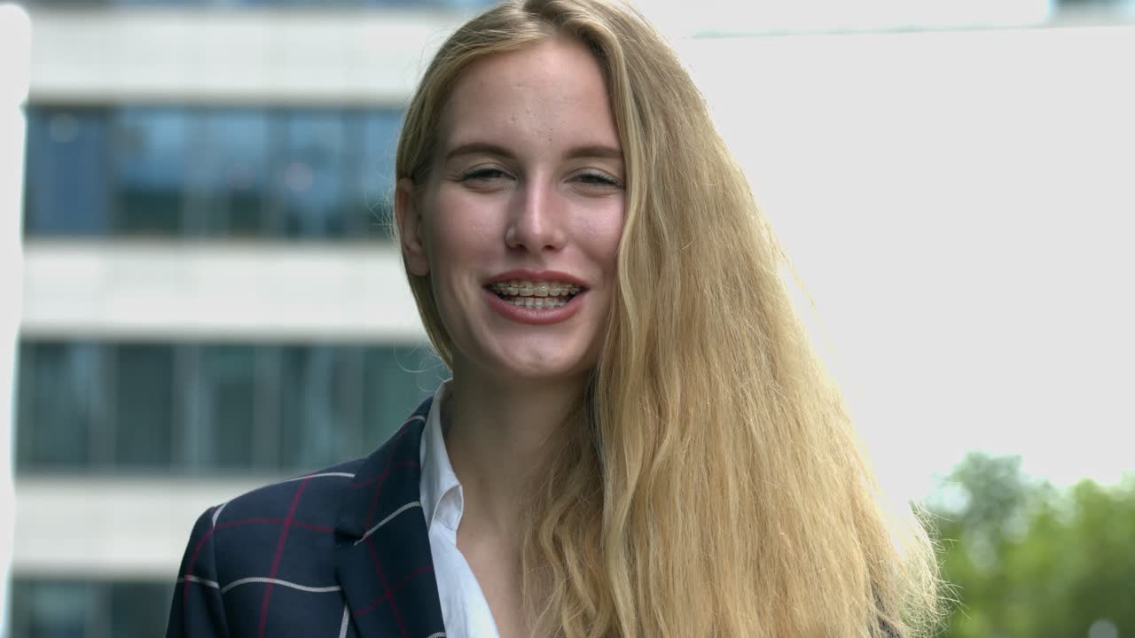 A young blonde female with dental braces stands outdoors smiling and laughing confidently