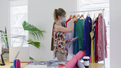 Female-fashion-designer-wearing-face-mask-looking-checking-on-dresses-at-studio