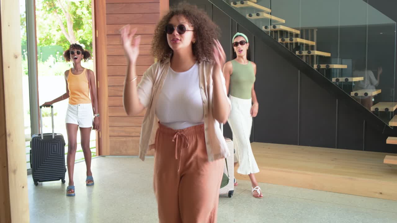 Mulheres afro-americanas chegando, a primeira mulher abrindo a porta da frente e puxando uma mala de rodinhas para o hall de entrada.