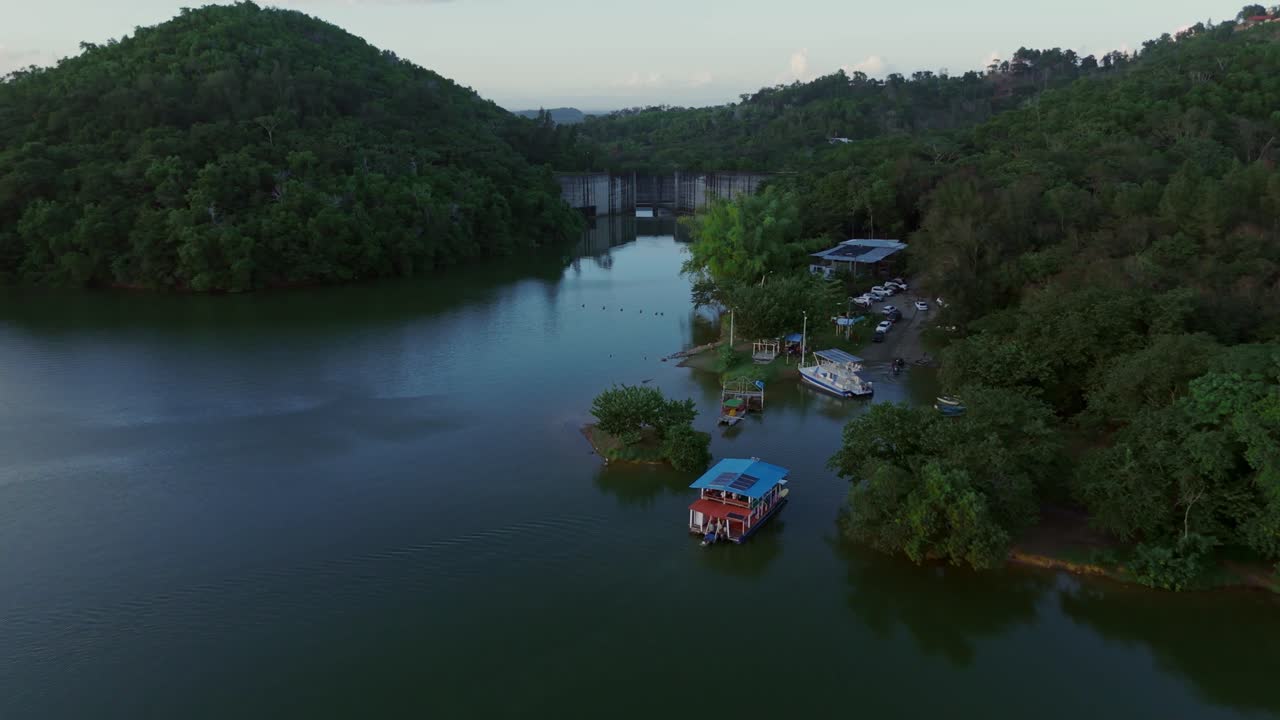 Ferry approaching dock at Hatillo Dam, Cotuí, Dominican Republic. Drone aerial view, dolly in flyover reveals dam wall