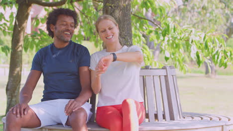 Couple-Wearing-Fitness-Clothing-Sit-On-Seat-Under-Tree-Checking-Activity-Monitor-On-Smartwatch