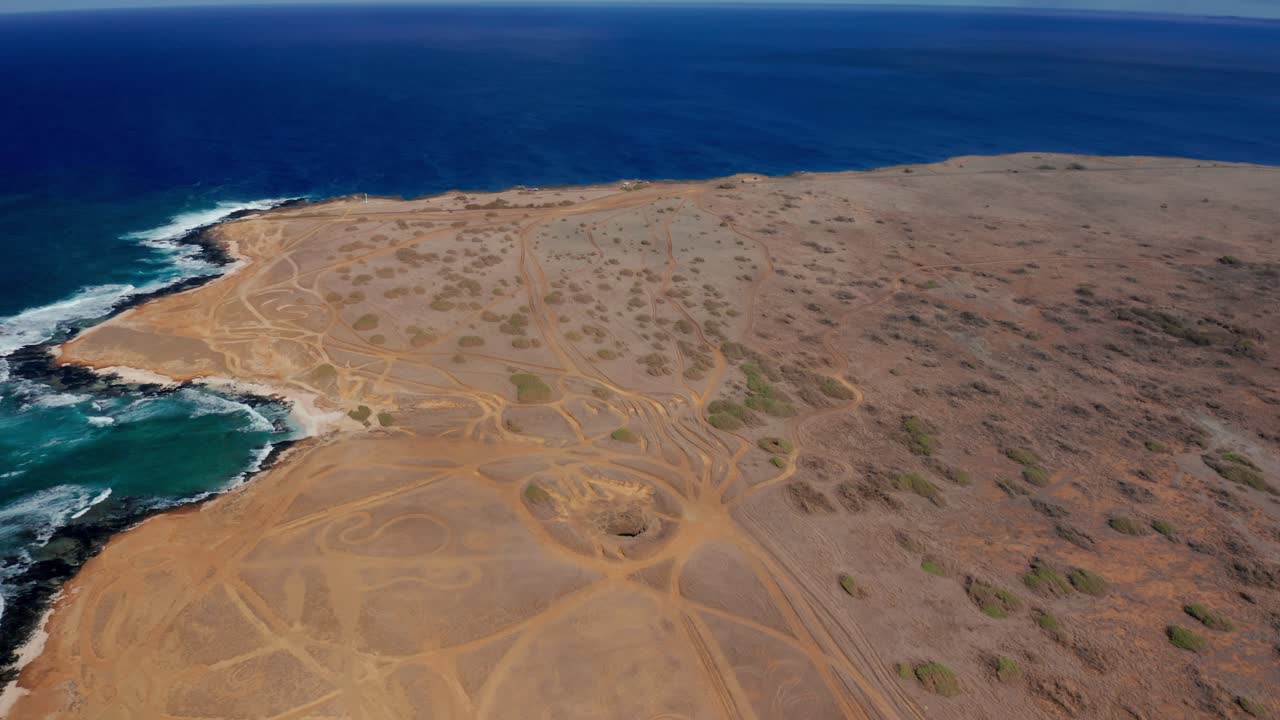 A drone drifts above a barren coastal plateau carved by looping off-road tracks as blue swells pound rock and lace the edge with foam, evoking remoteness and wind-scoured quiet