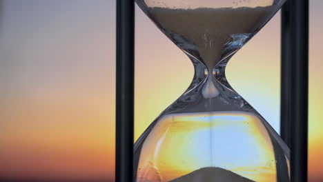 Fantasy-enchanted-hourglass-on-table,-sand-flowing-through-the-bulb-of-sandglass-on-orange-sky-background