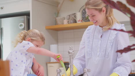 Madre-Usando-Guantes-De-Goma-En-Casa-En-La-Cocina-Con-Su-Hija-Pequeña-Divirtiéndose-Mientras-Lavan-En-El-Fregadero,-Filmada-En-Cámara-Lenta