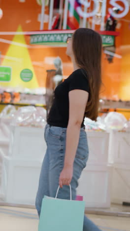 vista lateral de una mujer elegante con una camiseta negra y pantalones vaqueros que lleva una bolsa de compras mientras camina por delante de una tienda bien iluminada con paredes naranjas vibrantes, artículos minoristas en exhibición