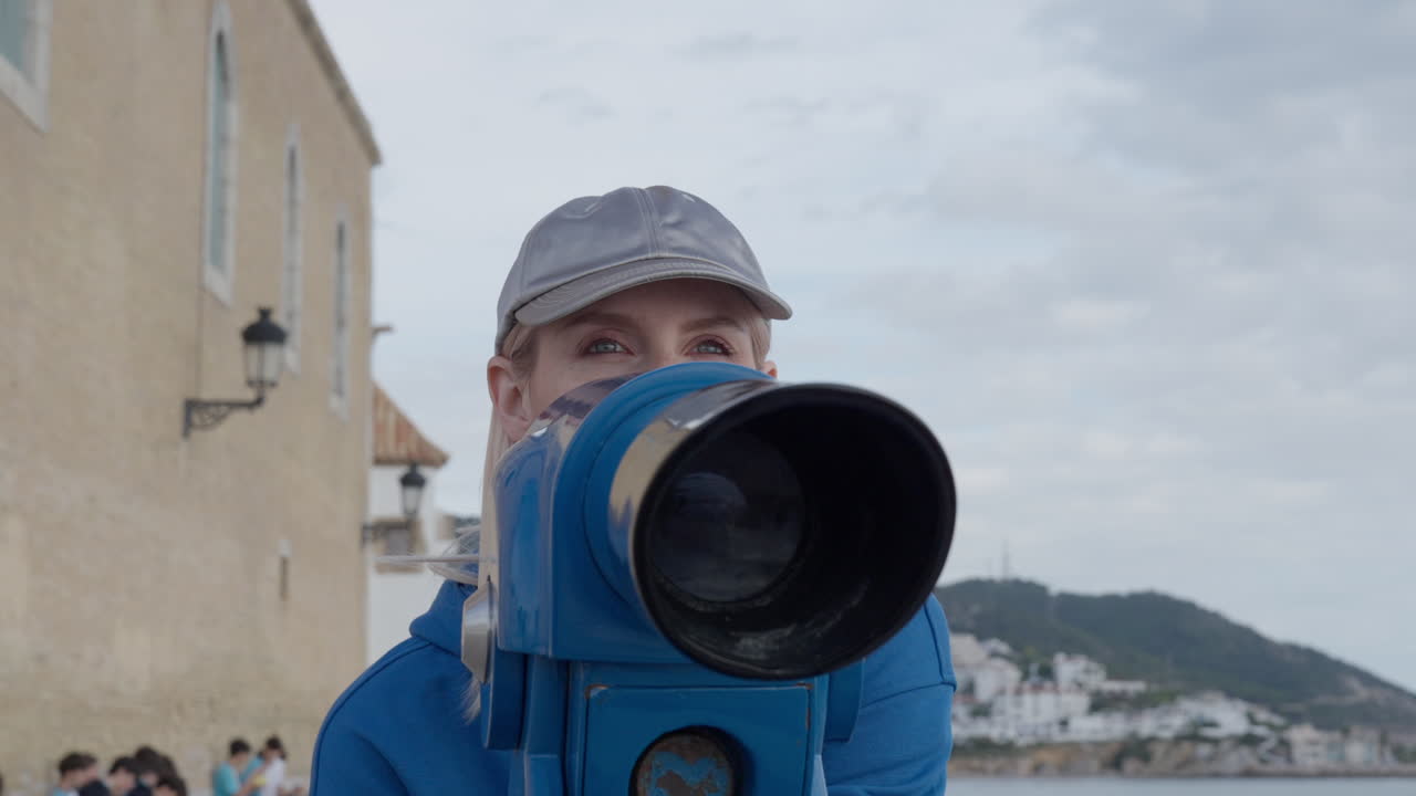 turista mujer blanca mirando a través de un telescopio azul, con una gorra deportiva con el fondo de la bahía del paisaje urbano nublado