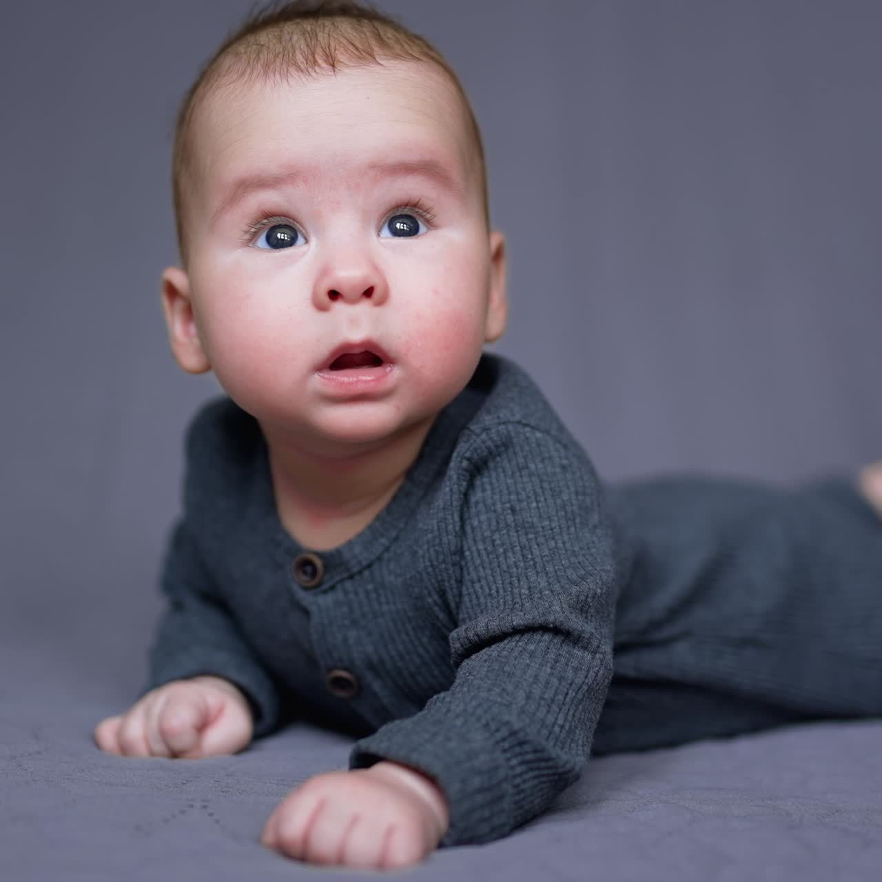 Sweet little boy in grey clothes lies on his belly. Healthy child looks around and then up. Grey blurred backdrop