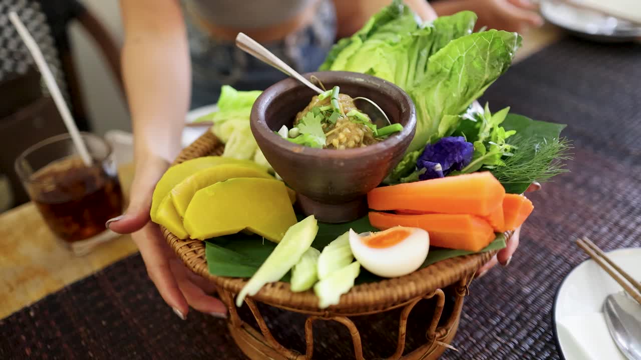 A person places a bamboo basket of Northern Thai roasted green chili dip, fresh vegetables, and boiled egg on a dining table in natural daylight