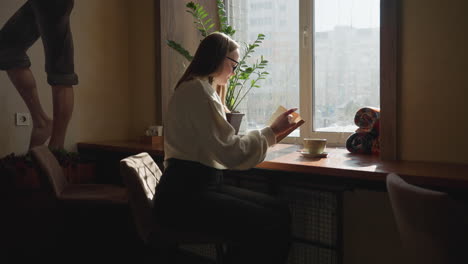 investor in white sweater focused on reading as she flips page of book beside sunlit window with city buildings in background, potted plant on wooden table and wall art