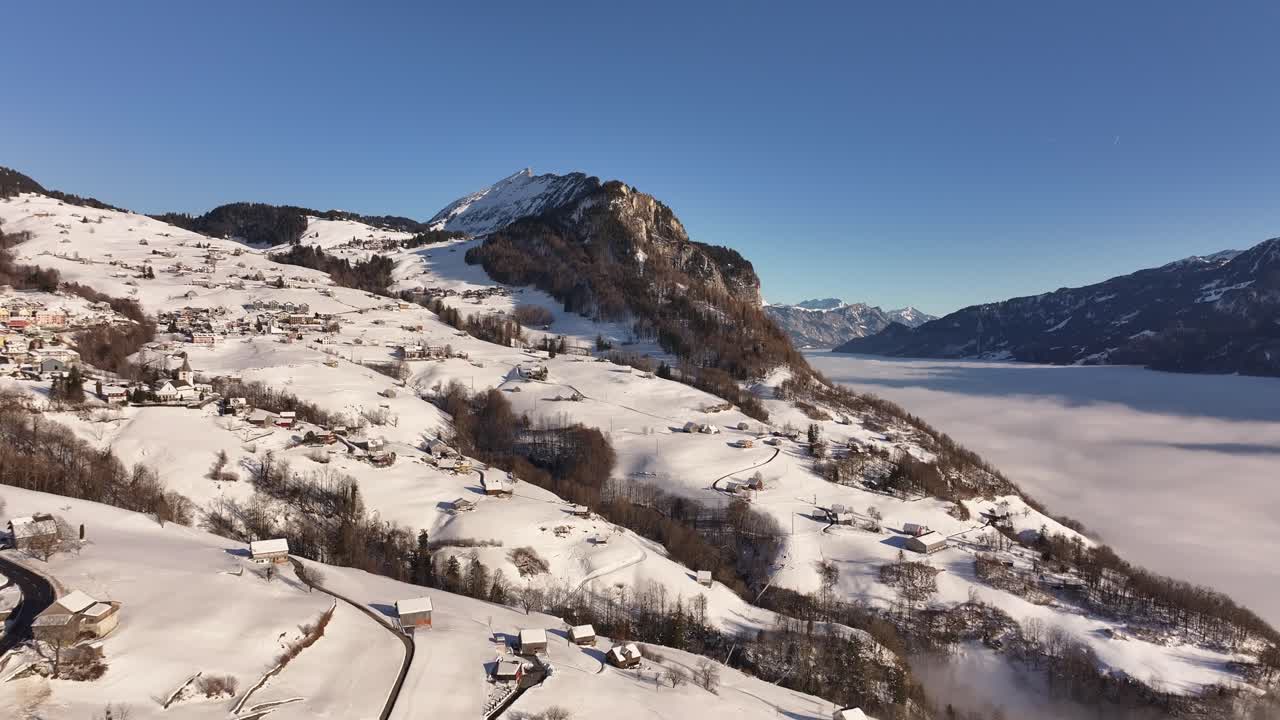 Aerial view of Amden in Glarus, Switzerland. Snow-covered alpine slopes meet a sea of fog blanketing the valley, with rugged mountain peaks under a clear blue sky.
