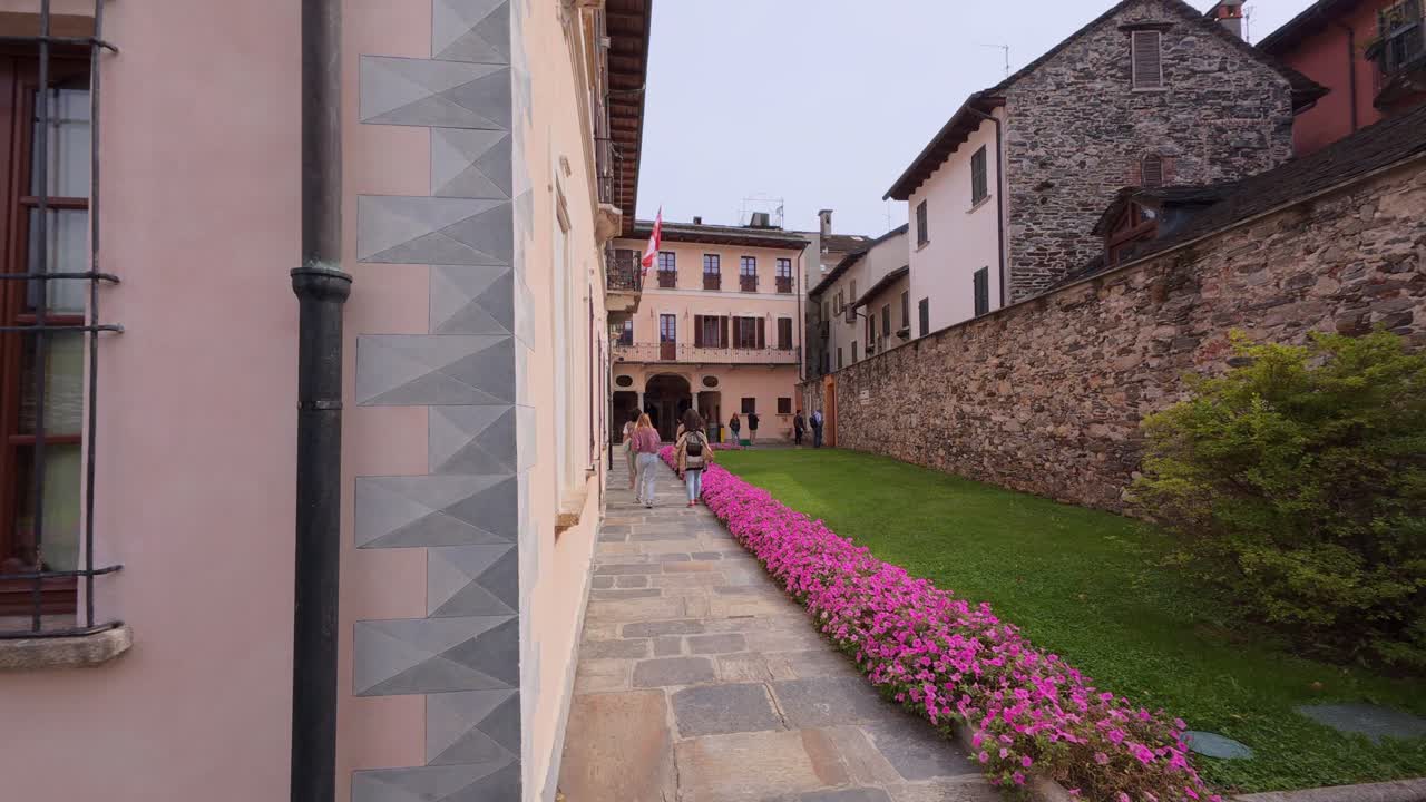 Charming alleyway in Orta San Giulio, Italy, with lush greenery and colorful flowers leading to a historic building