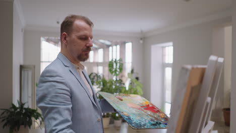 man takes art lesson at easel in studio. focused mature artist draws picture on canvas in workshop. bearded creator holds palette with pigments working