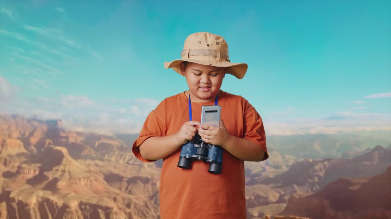Asian Boy With A Hat And Binoculars Looking At Smartphone And Smiling While Traveling At The Top Of Mountain. Boy Researcher Examines Something, Travel Tourism Adventure Concept