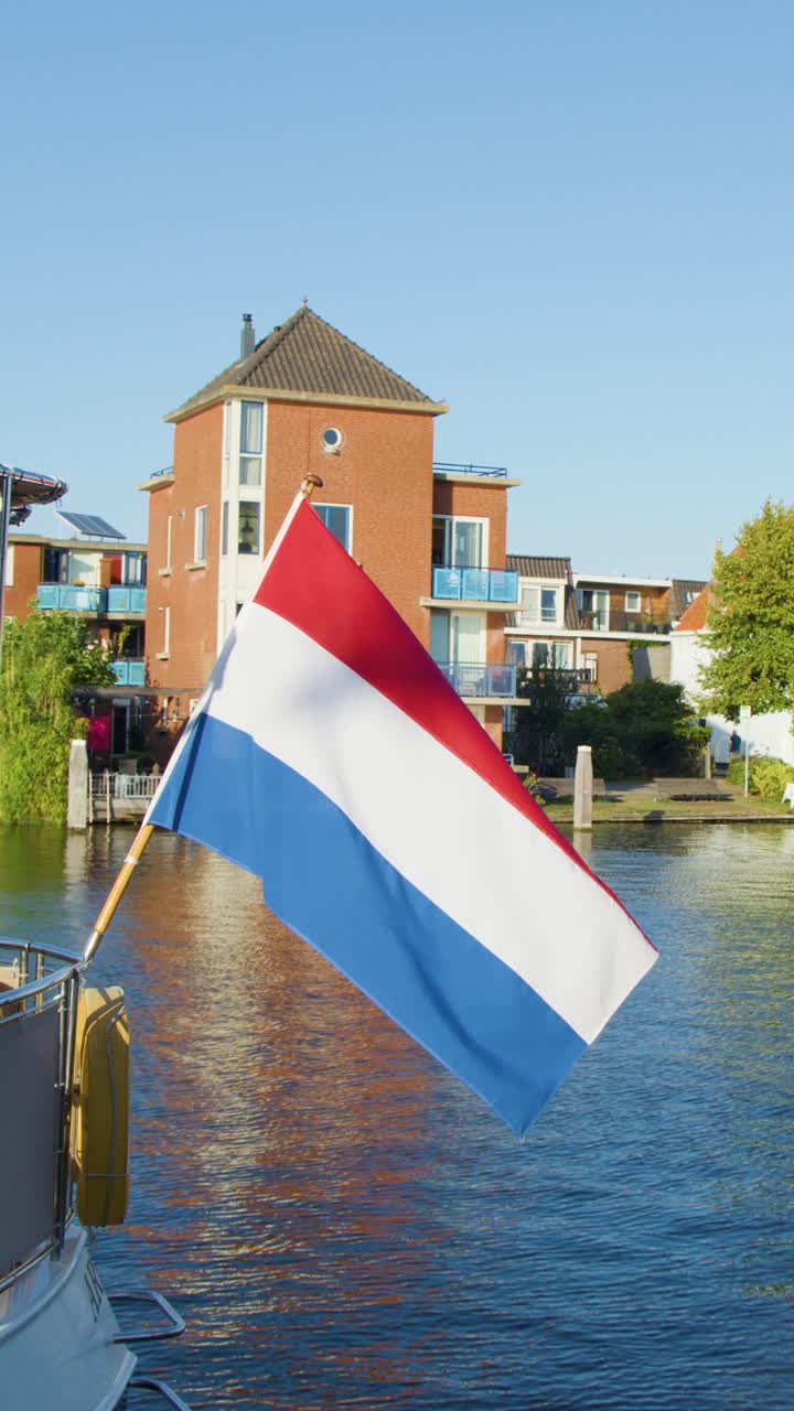 Niederländische Flagge weht auf einem Boot entlang des Haarlemer Kanals, historische Backsteingebäude und ruhiges Wasser, Tageslicht.