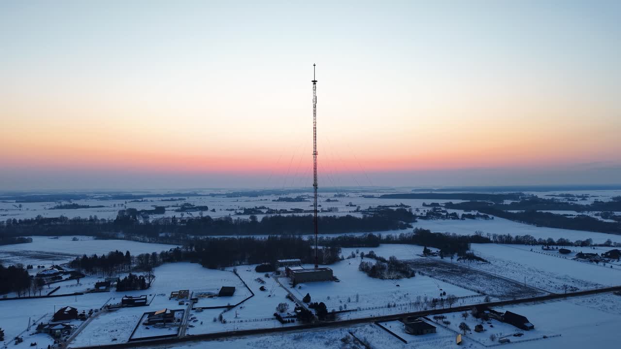 Riprese aeree di un paesaggio innevato con un'alta torre radio come fulcro al tramonto. L'ambiente invernale sereno è evidenziato dalle calde sfumature nel cielo.