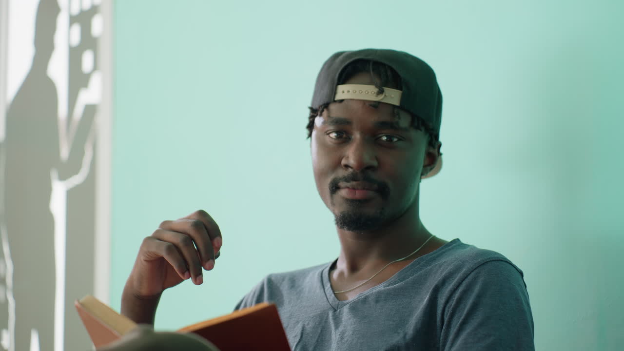 Close up of remote worker wearing cap looking up from reading with warm smile as bright sunlight streams through window casting shadow silhouette on teal wall in calm indoor setting