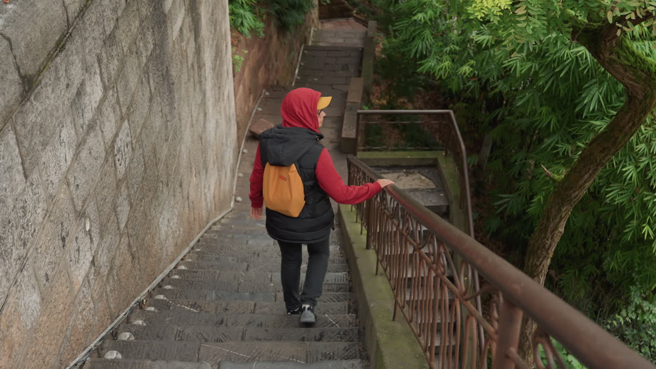 Pemandangan belakang seorang pelancong menuruni tangga sempit, tangan menyusuri pegangan tangan berkarat, hoodie merah dan tas kuning terlihat, gang berdaun dan dinding batu membingkai langkah lambat, hati-hati, fotografer perkotaan.