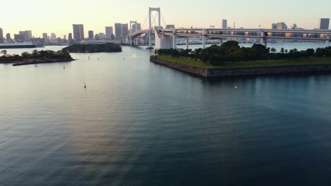 aerial drone flying low over water revealing rainbow bridge in odaiba tokyo city during sunset