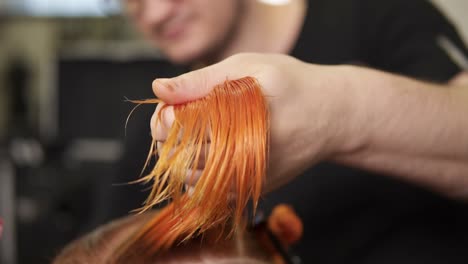 Stylish-male-hairdresser-cutting-woman's-hair-in-beauty-salon-while-holding-a-strand-of-hair-in-his-hand.-Young-woman-getting