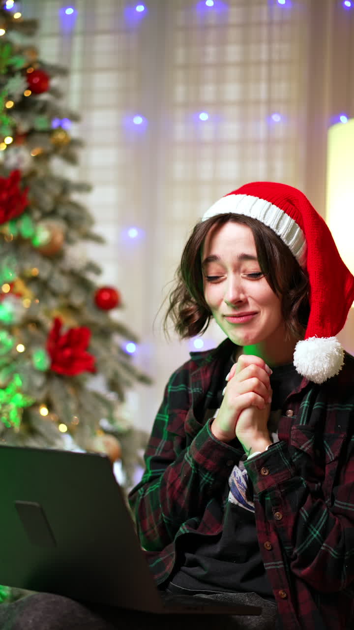 Happy rejoicing brunette girl looking at her laptop. Lady in Santa cap claps her hands excitedly. Christmas tree at backdrop. Vertical video.