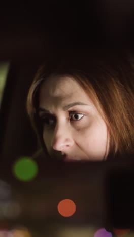vista en primera persona de una mujer ajustando el espejo del coche por la noche, con su rostro claramente reflejado en el espejo. las luces borrosas de la ciudad añaden una atmósfera de ensueño. tomada con una cámara de mano