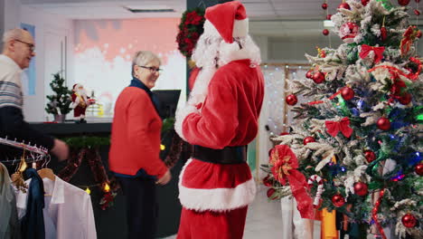 joyful african american worker dressed as santa claus in christmas ornate shopping mall clothing shop. employee jingling xmas bells and greeting happy clients in festive adorn fashion store
