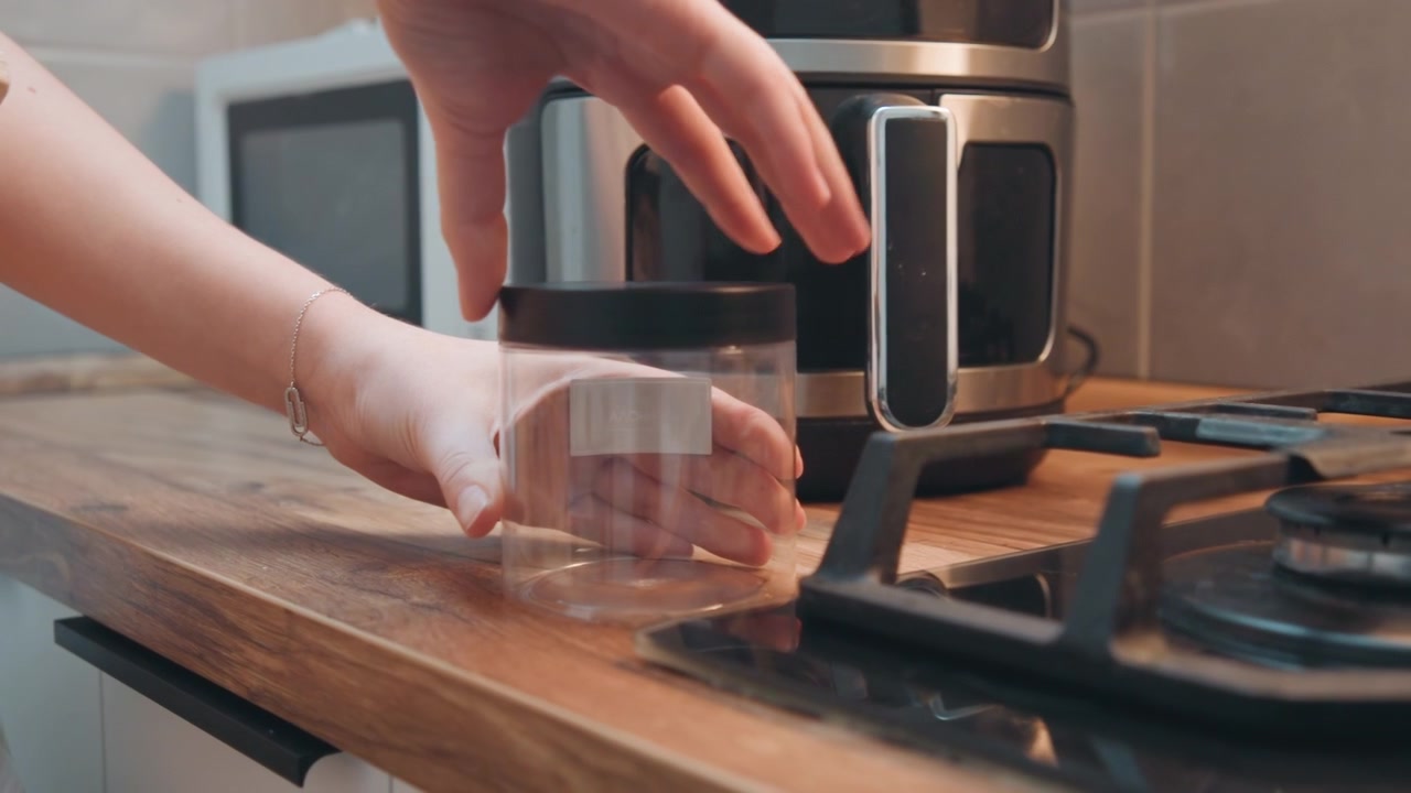 Barattolo sigillato su un piano di lavoro in legno in cucina. Primo piano delle mani che stringono il coperchio, accanto a una friggitrice ad aria e un fornello. L'attenzione è sulla conservazione ermetica di spezie e ingredienti, in un'atmosfera domestica accogliente con luce serale.