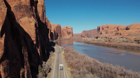 A-tracking-drone-shot-of-a-truck-driving-between-the-steep-cliffs,-known-as-the-“Wall-Street-Climbing-area”,-and-the-Colorado-River,-near-Moab,-Utah