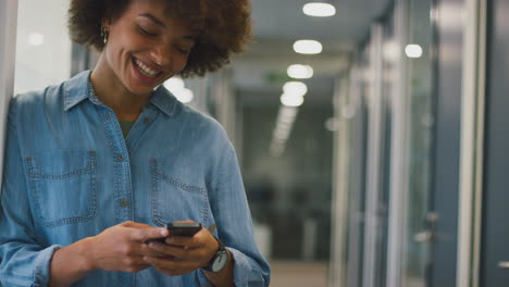 smiling businesswoman standing in corridor of modern office texting with mobile phone