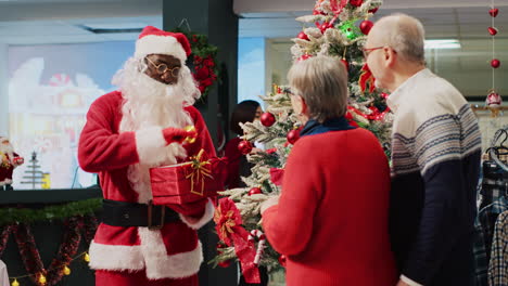 Trabajador-Afroamericano-Vestido-Como-Papá-Noel-Tocando-La-Campana-Y-Sosteniendo-Un-Regalo-De-Navidad-Junto-A-Un-árbol-De-Navidad-Bellamente-Decorado,-Hablando-Con-Una-Pareja-Mayor-Comprando-En-Una-Tienda-De-Moda-Decorada-Con-Navidad