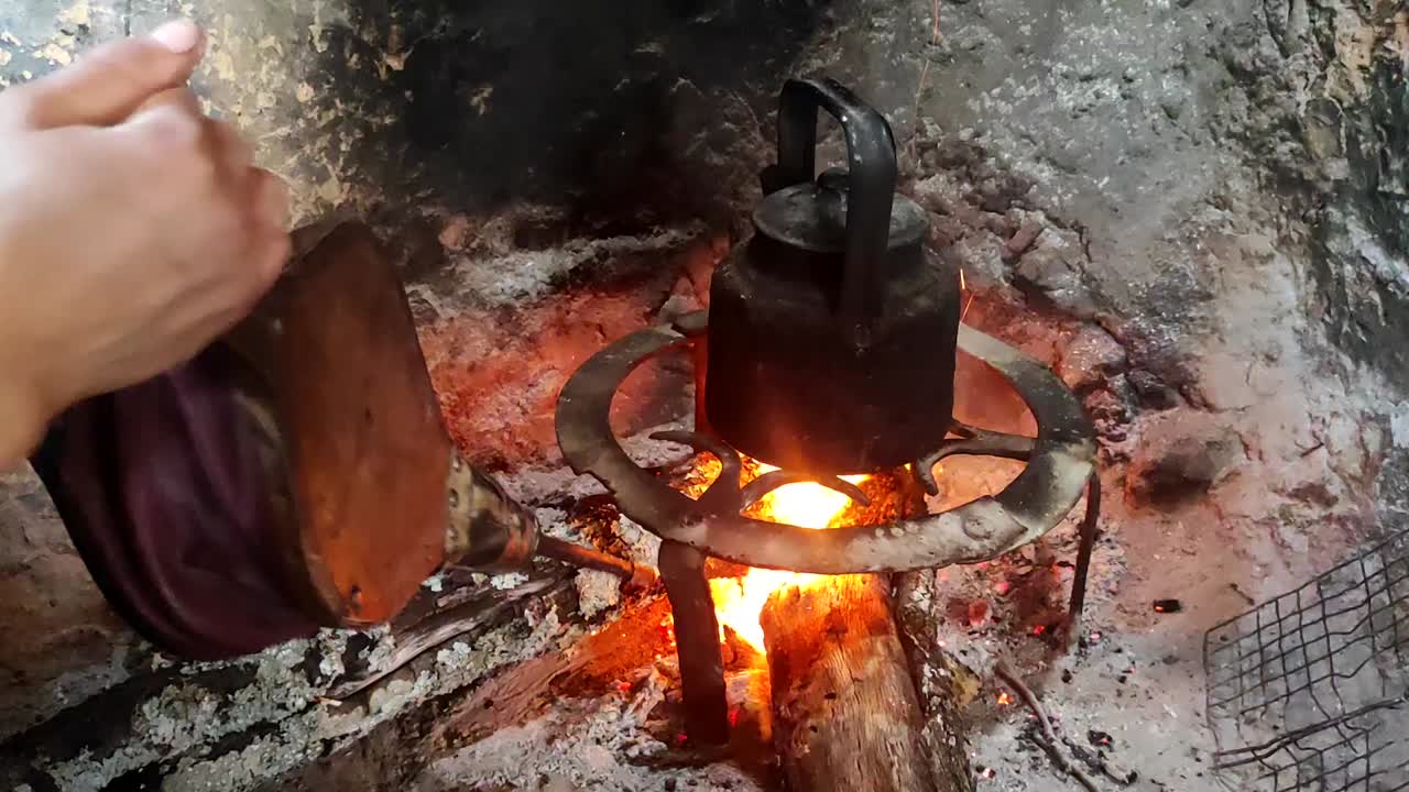 primer plano de cocinar la cena en una olla sobre un fuego de madera en una chimenea de ladrillo en una cocina tradicional y manos de mujer soplando las brasas con un fuelle