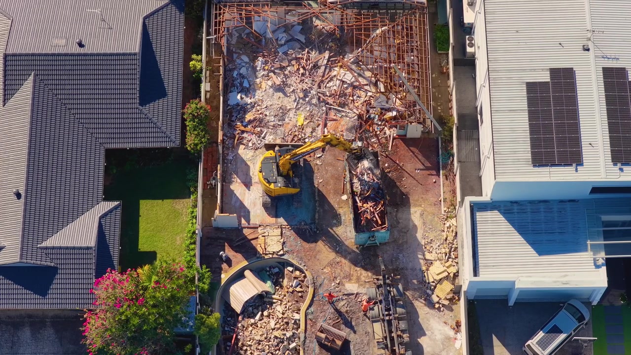 Top-down drone shot of heavy machinery loading debris into a skip bin during house demolition.