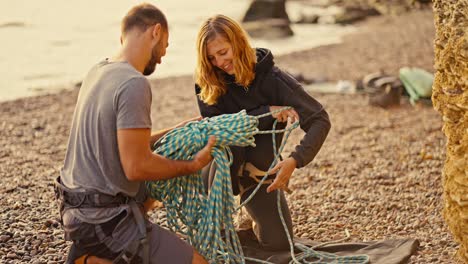 A-blonde-girl-in-a-black-summer-uniform,-together-with-her-brunette-boyfriend-in-a-gray-T-shirt,-takes-out-a-blue-rope-and-unravels-it-in-order-to-set-up-a-belay-and-start-rock-climbing-on-a-rocky-seashore