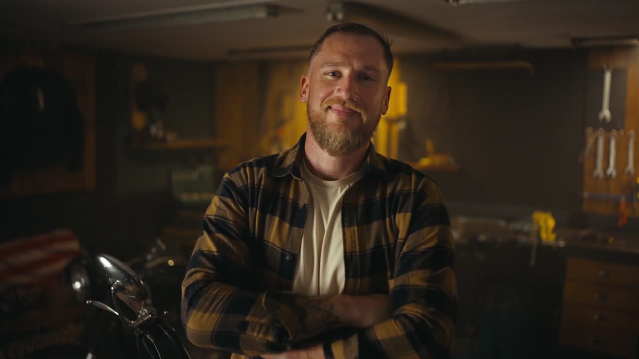 Portrait of a happy blond guy with a beard in a checkered shirt who crossed his arms over his chest and poses in his workshop