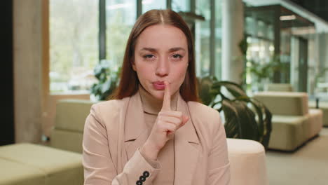 portrait of serious young caucasian businesswoman in formals making silence gesture in office lobby