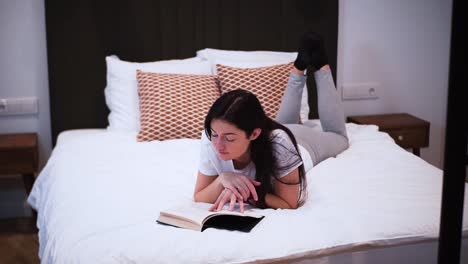 a young woman is reading a book with black hardcover in her bedroom, laying on the bed, she spends her free time with a book, relaxing at home, turns the page, flips the page, close-up shot, smiling