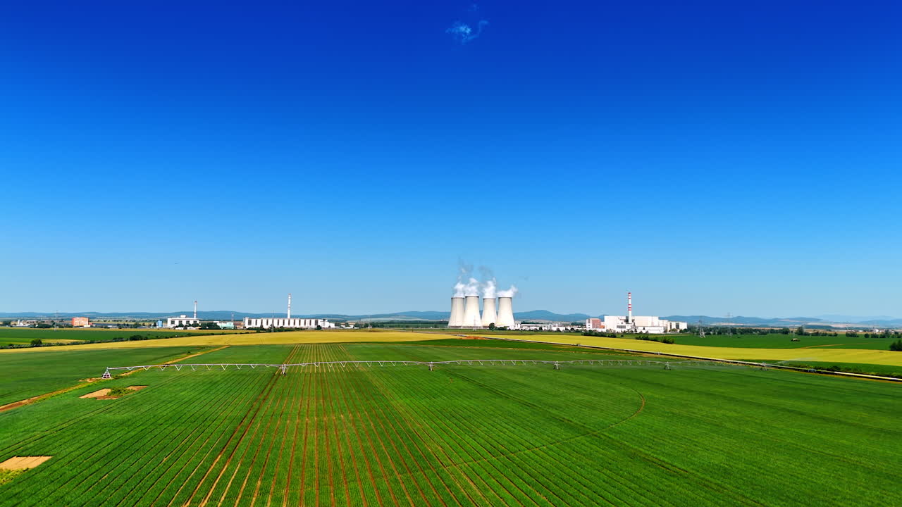 Power plant by green fields. A large power plant with cooling towers is seen next to vibrant green fields under a clear blue sky during daytime