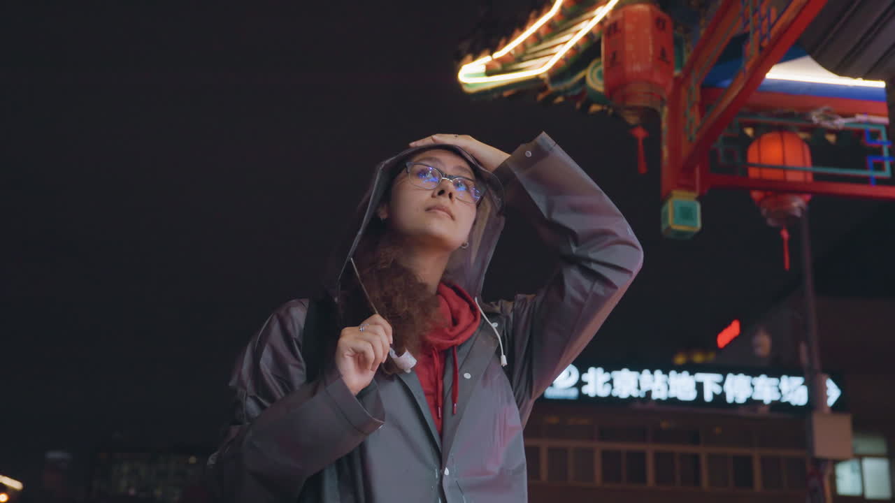 Notte sotto un baldacchino di lanterne, donna asiatica che guarda in alto con meraviglia, uno zaino in spalla incappucciato che si sistema il cappotto, tetto in stile tempio illuminato al neon, bokeh cinematografico e segnaletica, pausa del viaggiatore e curiosità culturale