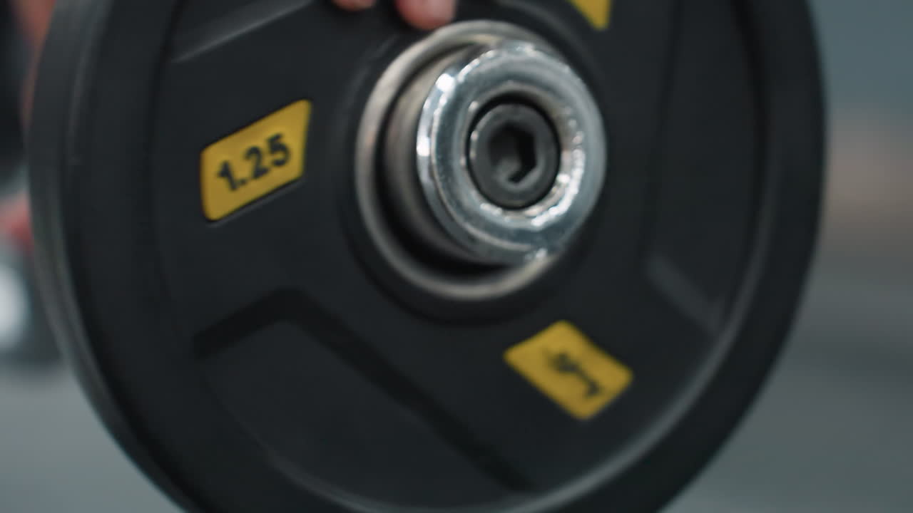 Close up action of person inserting 1.25 weight plate onto barbell in gym environment, with blurred view of clip on rubber floor, symbolizing workout preparation and strength training routine indoors