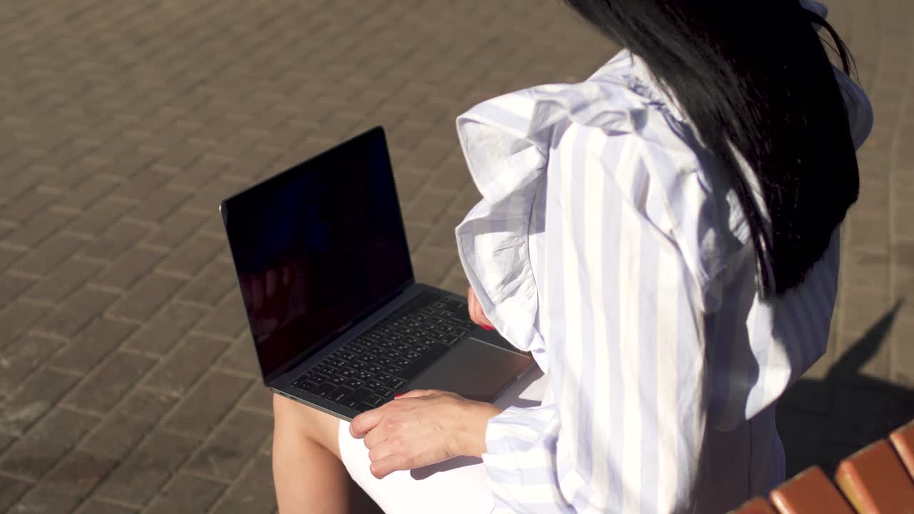 Brunete typing on laptop sitting on a bench in park