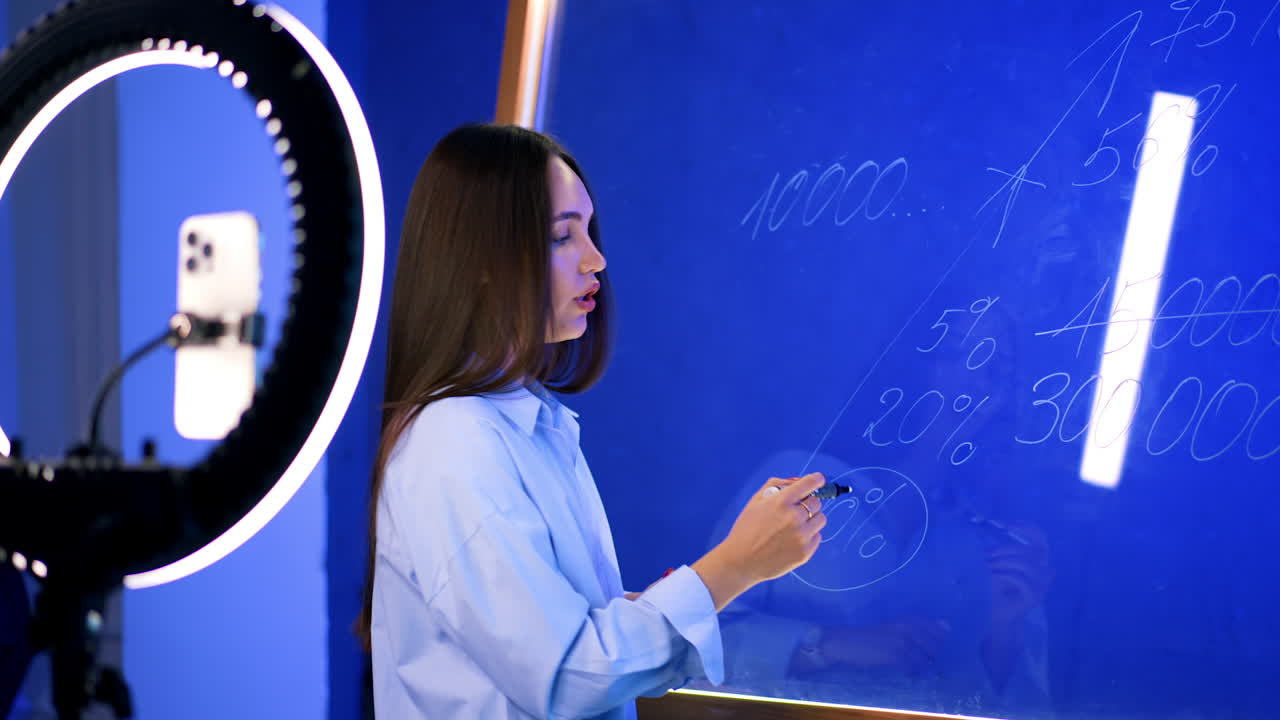 Brunette speaks to camera and circles the numbers on the glass wall. Recording video for frog or streaming.