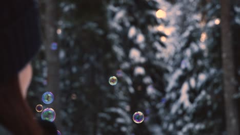 brunette woman with winter clothes blowing rainbow colored soap bubbles flying suspended in the air, with a snowy forest in the background