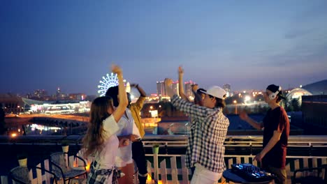 multiracial group of friends is dancing on rooftop at outdoor party with dj working with equipment. modern city nightlife, youth and entertainment concept.