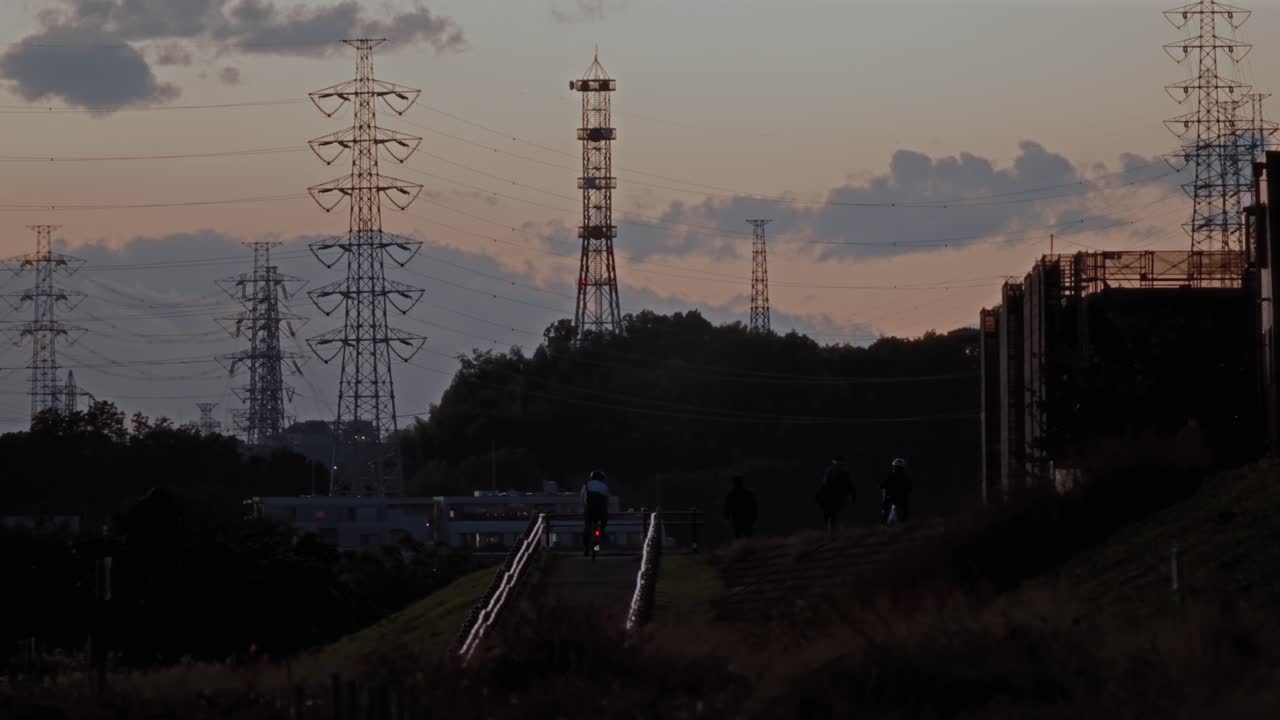 電線や鉄塔のそばを自転車で通り過ぎるサイクリストがいる静かな夕暮れの風景。日常生活、都市インフラ、穏やかな夜の雰囲気を捉えている。