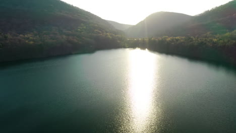 Cinematic-aerial-pull-away-over-forest-lake-surrounded-by-green-mountains-at-sunset