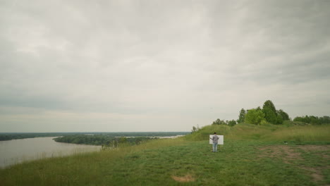 a painter wearing a hat and checkered shirt is intently focused on creating art at an easel in a vibrant green field under a cloudy sky. in the background, a serene lake stretches across the landscape