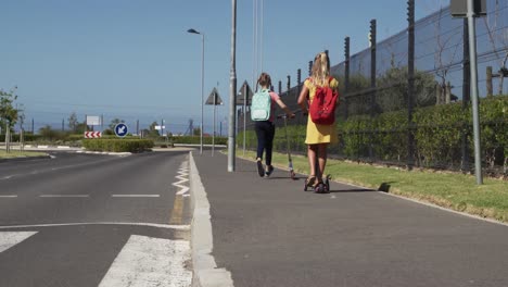 two girls with school bags riding scooters on footpath